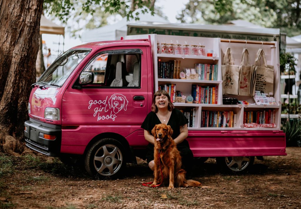 Photo of Good Girl Books owner Rayann Streeter and Poppy, OG Good Girl in front of the pink Mitsubishi Bookmobile