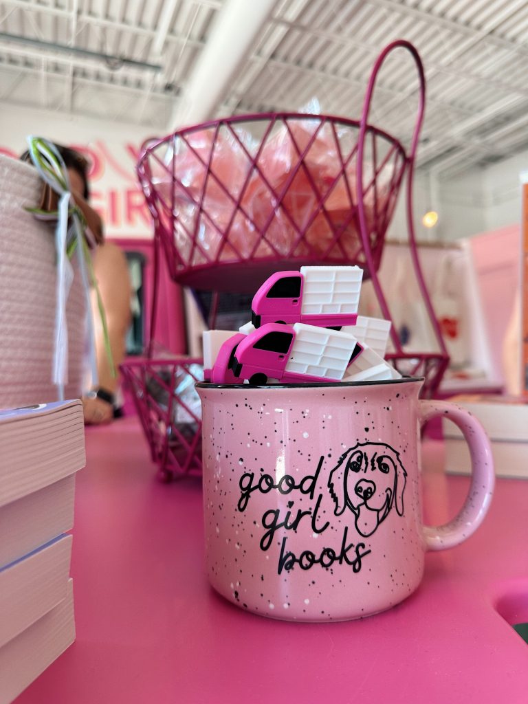 Pink ceramic mug that reads 'good girl books' with a golden retriver doodle on a pink table with plastic bookmarks in the shape of Little Miss Smuttie, the pink book truck