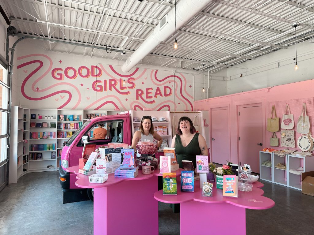 Two women stand in a large white & pink painted bookstore in front of Little Miss Smuttie, the pink book truck